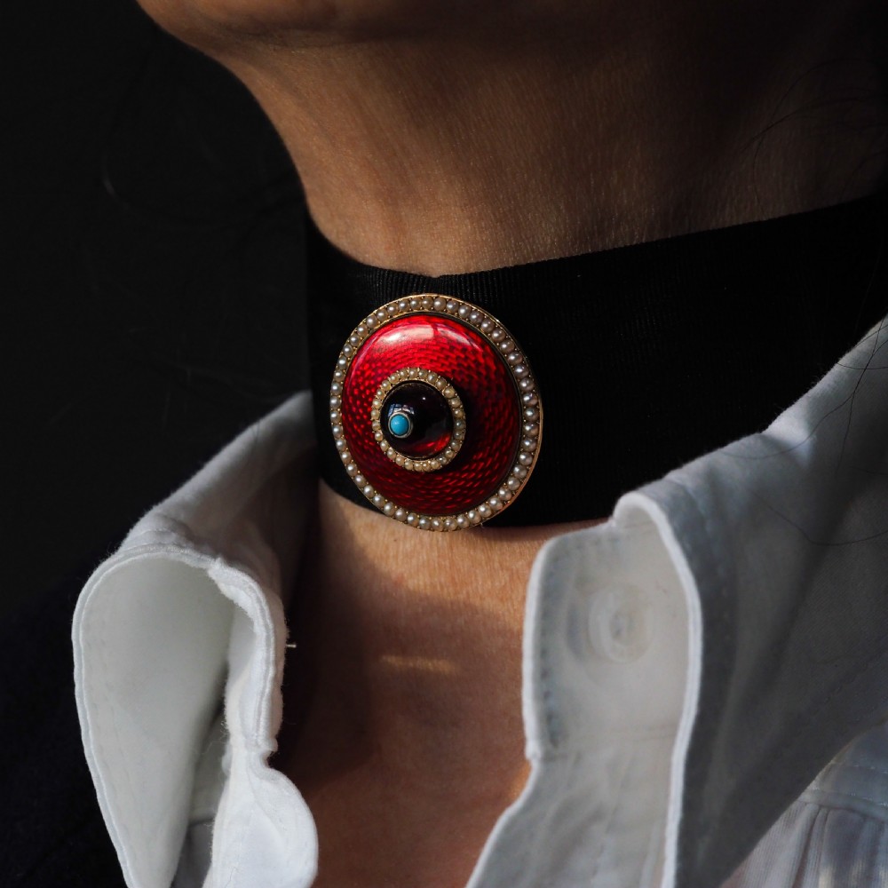 A circular Victorian red guilloche enamel brooch featuring concentric seed pearl borders, a central dark garnet cabochon, and a small turquoise accent set in gold. View V