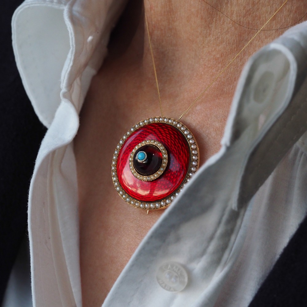 A circular Victorian red guilloche enamel brooch featuring concentric seed pearl borders, a central dark garnet cabochon, and a small turquoise accent set in gold. View VI