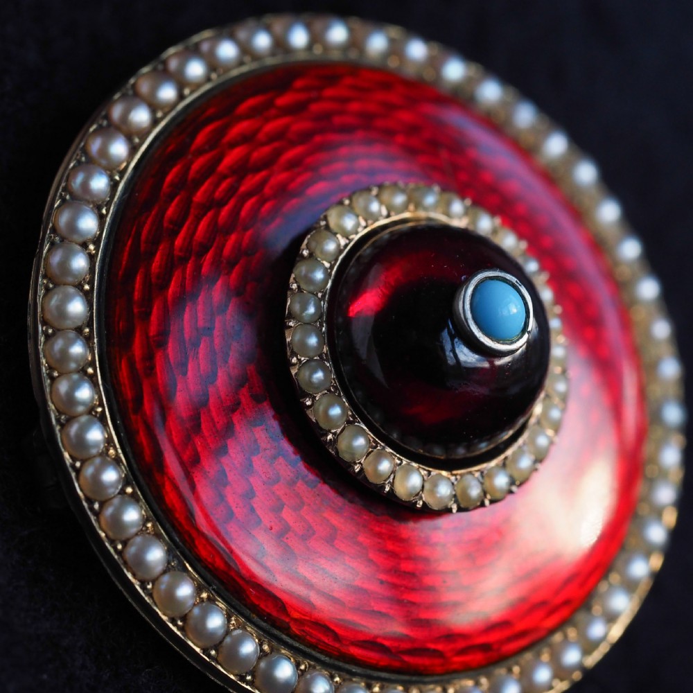 A circular Victorian red guilloche enamel brooch featuring concentric seed pearl borders, a central dark garnet cabochon, and a small turquoise accent set in gold. View VII