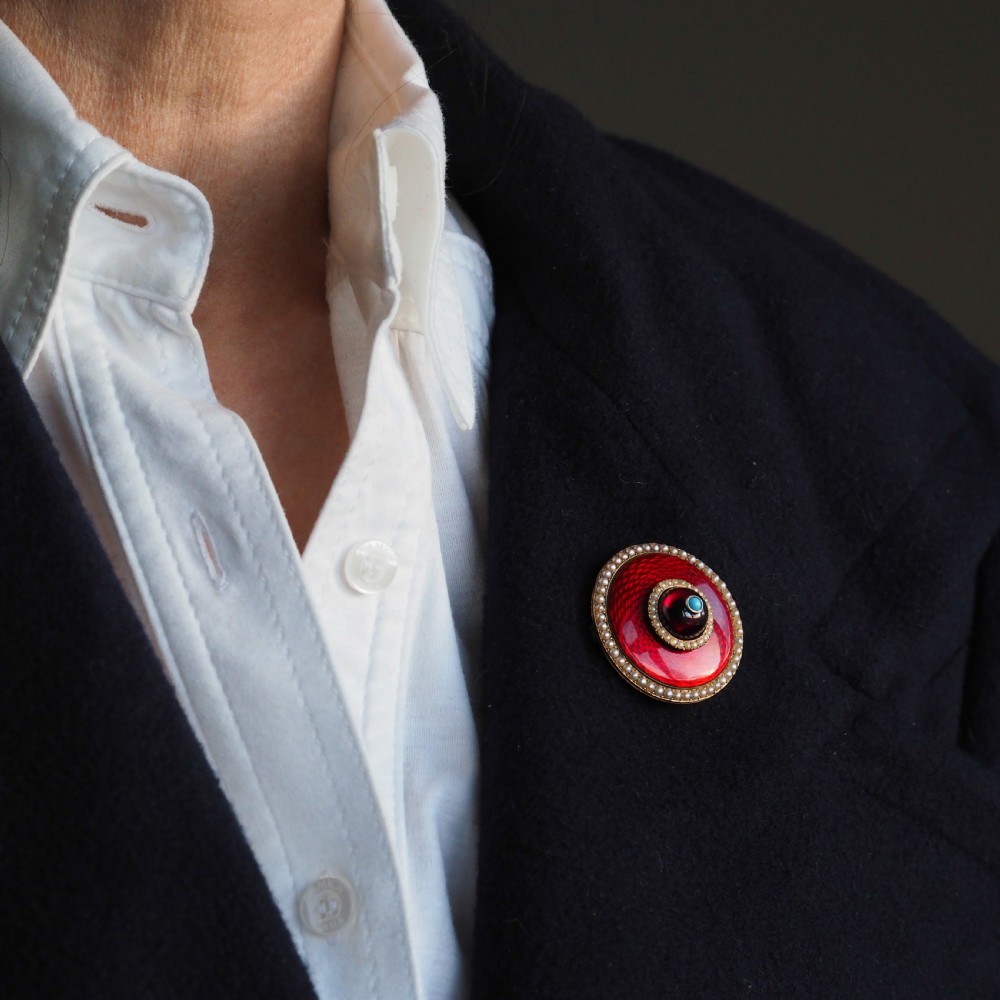 A circular Victorian red guilloche enamel brooch featuring concentric seed pearl borders, a central dark garnet cabochon, and a small turquoise accent set in gold. View VIII