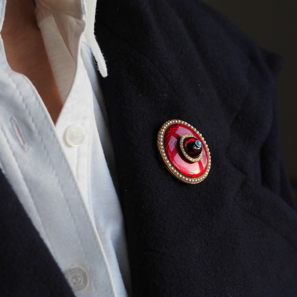 A circular Victorian red guilloche enamel brooch featuring concentric seed pearl borders, a central dark garnet cabochon, and a small turquoise accent set in gold. View IX