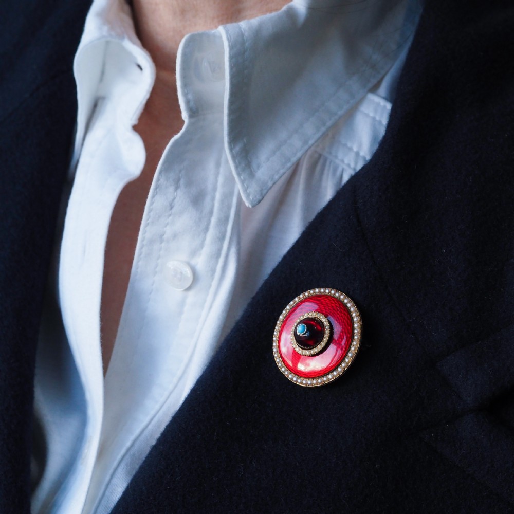 A circular Victorian red guilloche enamel brooch featuring concentric seed pearl borders, a central dark garnet cabochon, and a small turquoise accent set in gold. View X