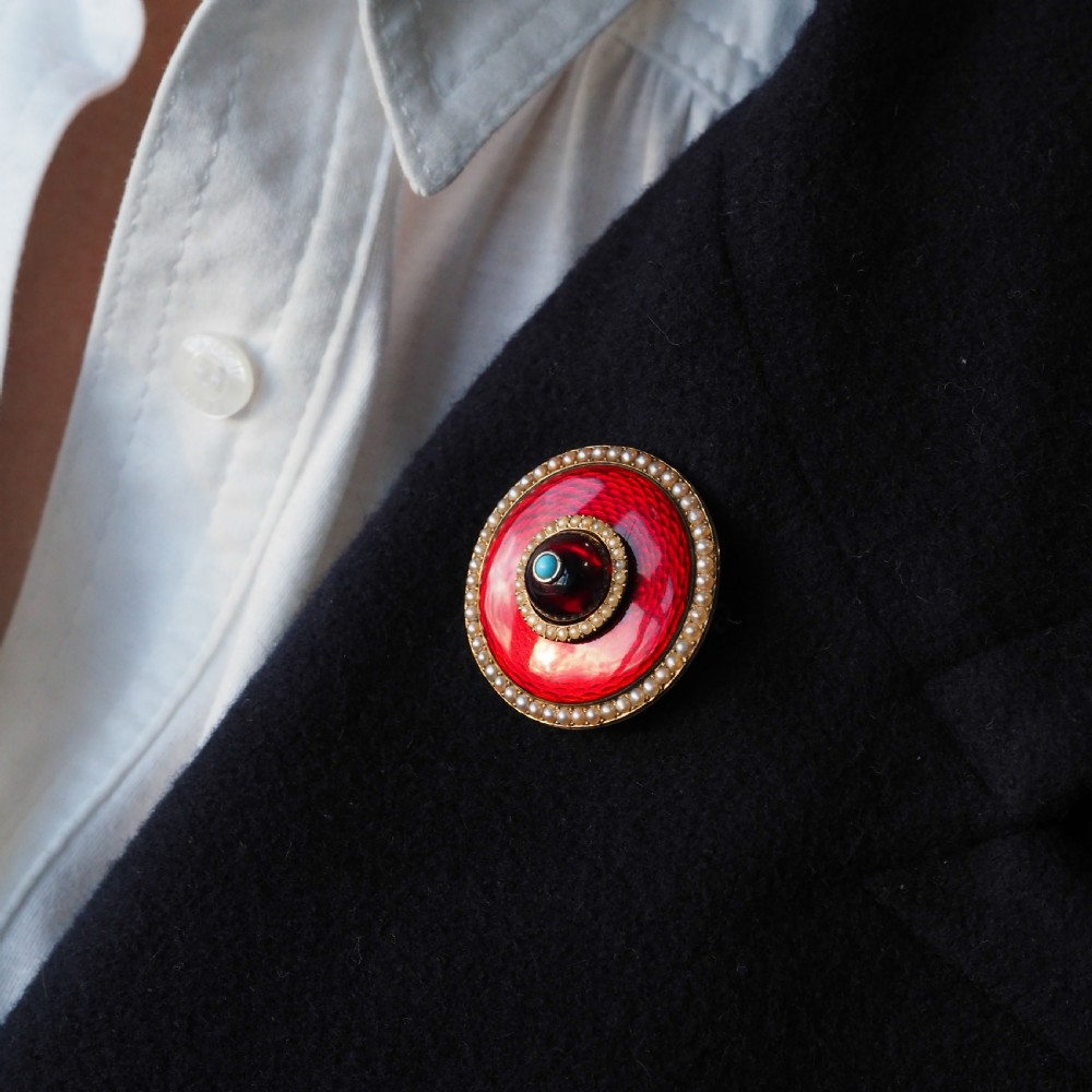A circular Victorian red guilloche enamel brooch featuring concentric seed pearl borders, a central dark garnet cabochon, and a small turquoise accent set in gold. View XI