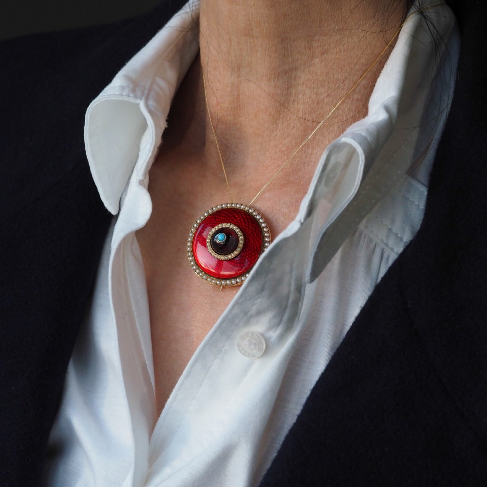 A circular Victorian red guilloche enamel brooch featuring concentric seed pearl borders, a central dark garnet cabochon, and a small turquoise accent set in gold. View XV