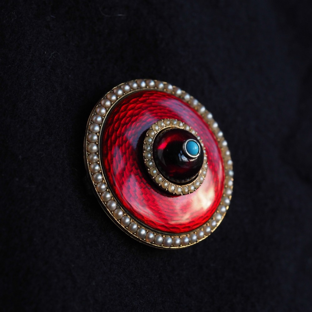 A circular Victorian red guilloche enamel brooch featuring concentric seed pearl borders, a central dark garnet cabochon, and a small turquoise accent set in gold. View XVII