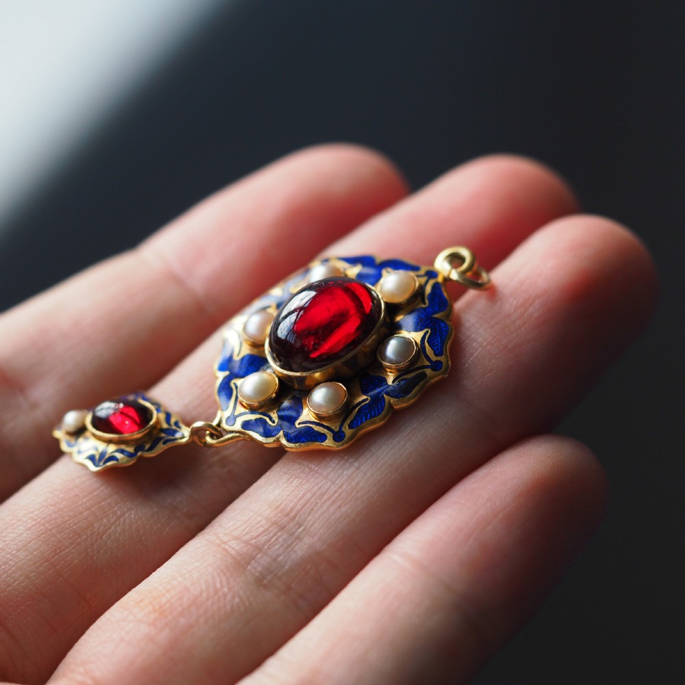 An 18k yellow gold Victorian Holbeinesque pendant featuring a central red cabochon garnet surrounded by pearls, blue enamel, and an articulated gemstone drop. View IV