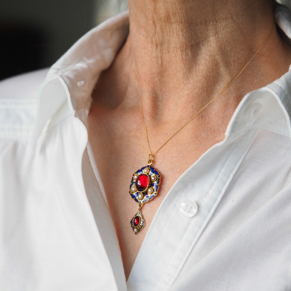 An 18k yellow gold Victorian Holbeinesque pendant featuring a central red cabochon garnet surrounded by pearls, blue enamel, and an articulated gemstone drop. View V
