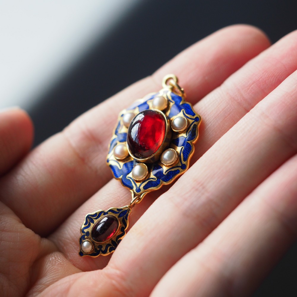 An 18k yellow gold Victorian Holbeinesque pendant featuring a central red cabochon garnet surrounded by pearls, blue enamel, and an articulated gemstone drop. View VI