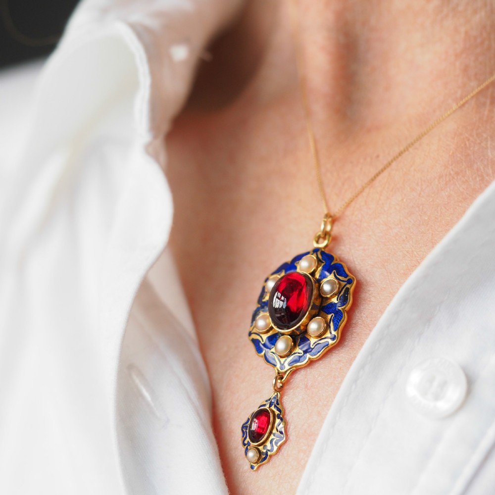 An 18k yellow gold Victorian Holbeinesque pendant featuring a central red cabochon garnet surrounded by pearls, blue enamel, and an articulated gemstone drop. View VII