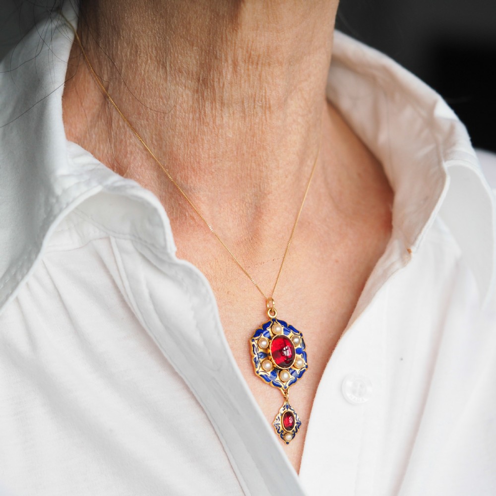 An 18k yellow gold Victorian Holbeinesque pendant featuring a central red cabochon garnet surrounded by pearls, blue enamel, and an articulated gemstone drop. View X