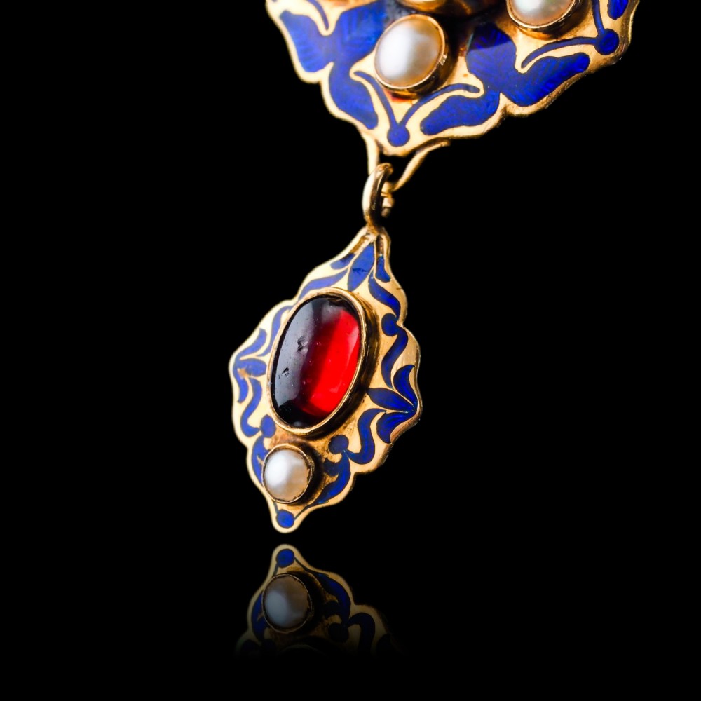 An 18k yellow gold Victorian Holbeinesque pendant featuring a central red cabochon garnet surrounded by pearls, blue enamel, and an articulated gemstone drop. View XVI