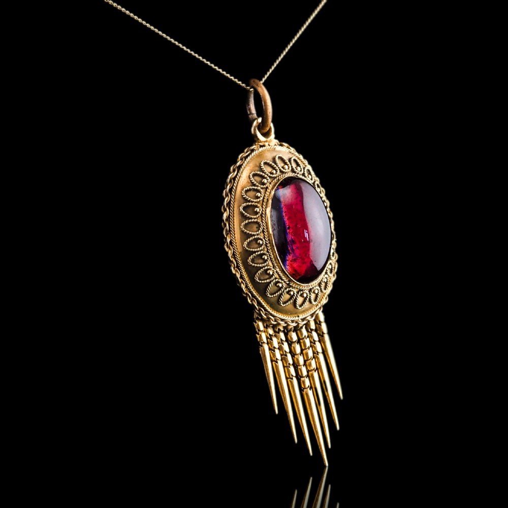 A Victorian 15ct gold oval Etruscan Revival pendant set with a central cabochon garnet and decorated with fine wirework and hanging spike tassels. View V