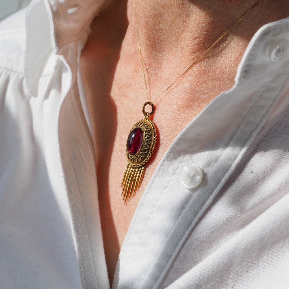 A Victorian 15ct gold oval Etruscan Revival pendant set with a central cabochon garnet and decorated with fine wirework and hanging spike tassels. View XIV