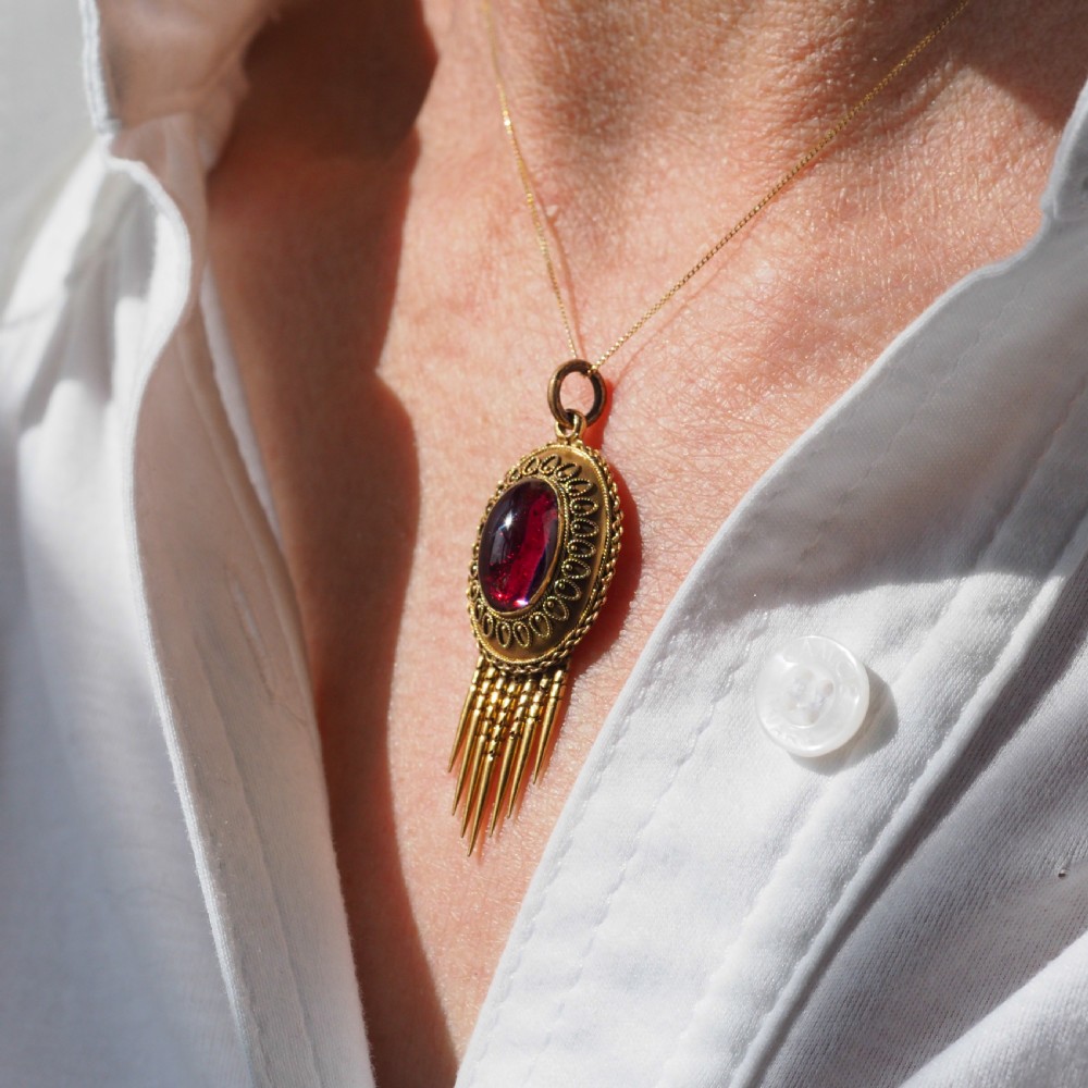 A Victorian 15ct gold oval Etruscan Revival pendant set with a central cabochon garnet and decorated with fine wirework and hanging spike tassels. View XV