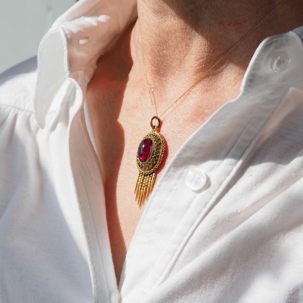 A Victorian 15ct gold oval Etruscan Revival pendant set with a central cabochon garnet and decorated with fine wirework and hanging spike tassels. View XVI