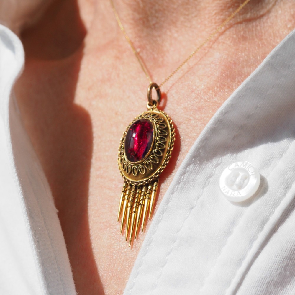 A Victorian 15ct gold oval Etruscan Revival pendant set with a central cabochon garnet and decorated with fine wirework and hanging spike tassels. View XVII