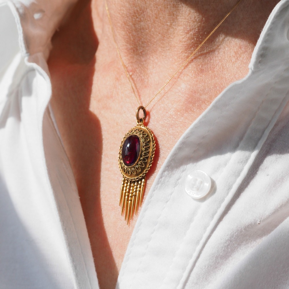 A Victorian 15ct gold oval Etruscan Revival pendant set with a central cabochon garnet and decorated with fine wirework and hanging spike tassels. View XVIII