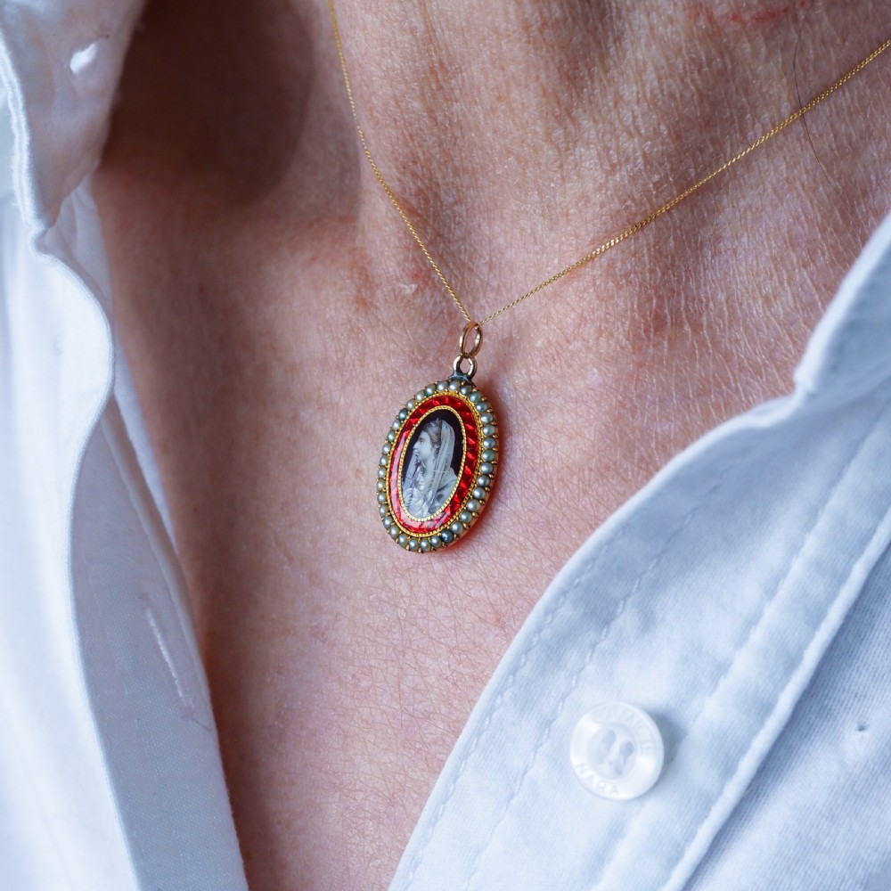 An oval Georgian gold pendant featuring a sepia miniature profile portrait surrounded by a red enamel border and a frame of seed pearls on a fine gold chain. View X