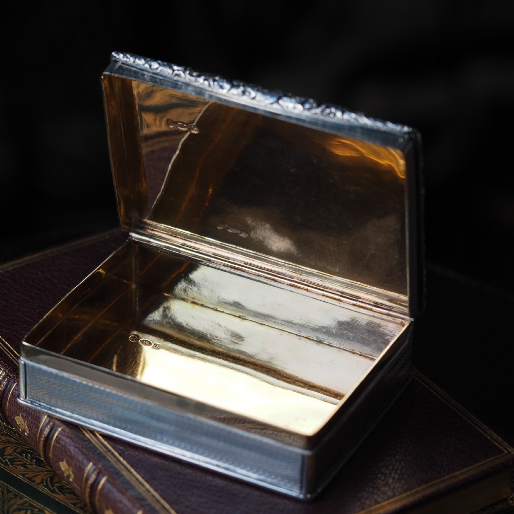 A rectangular sterling silver Georgian snuff box featuring a chased floral border, engine-turned detailing, and a central engraved crest. View XX