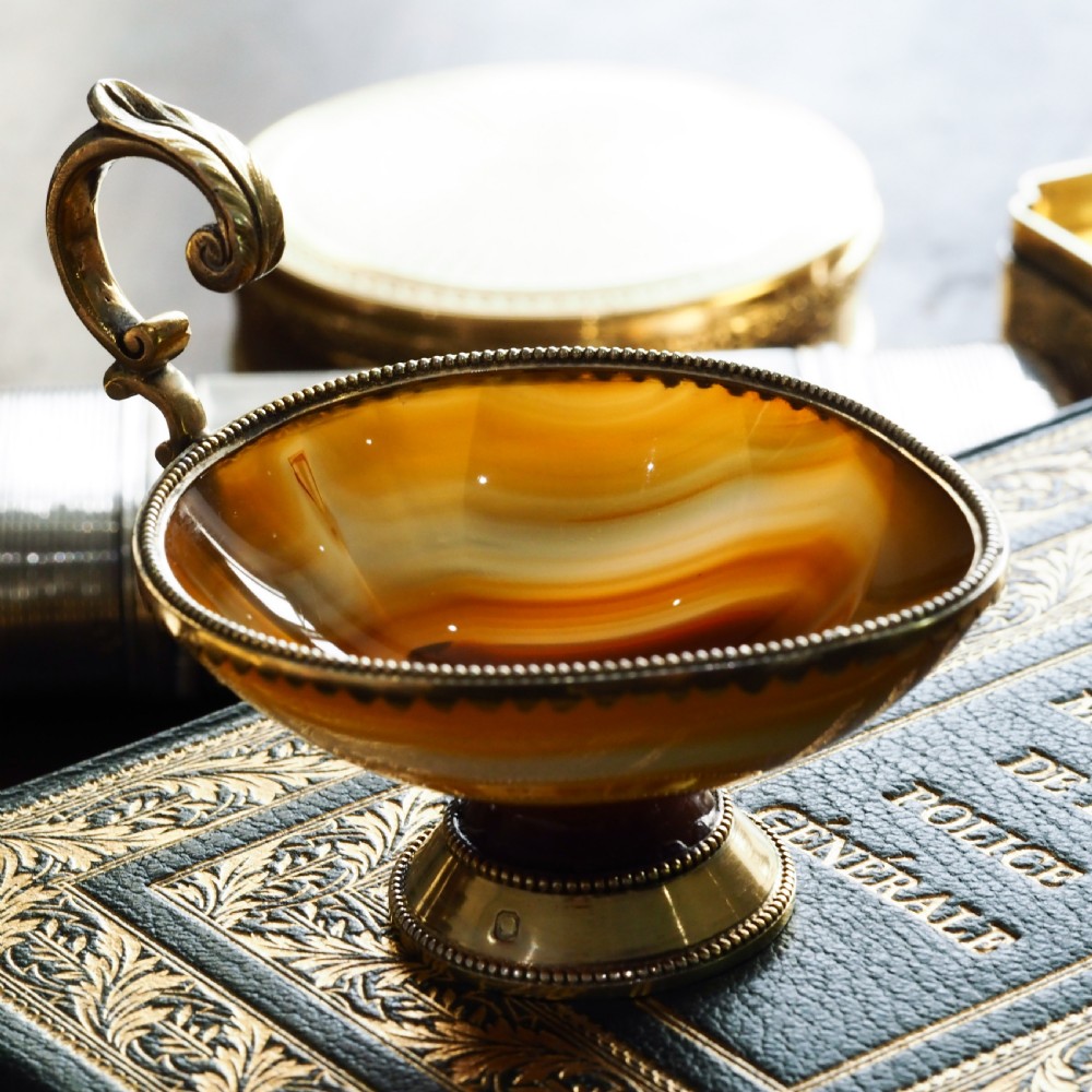 A 19th-century carved brown banded agate cup featuring a silver-gilt rim, pedestal base, and an ornate scroll handle.