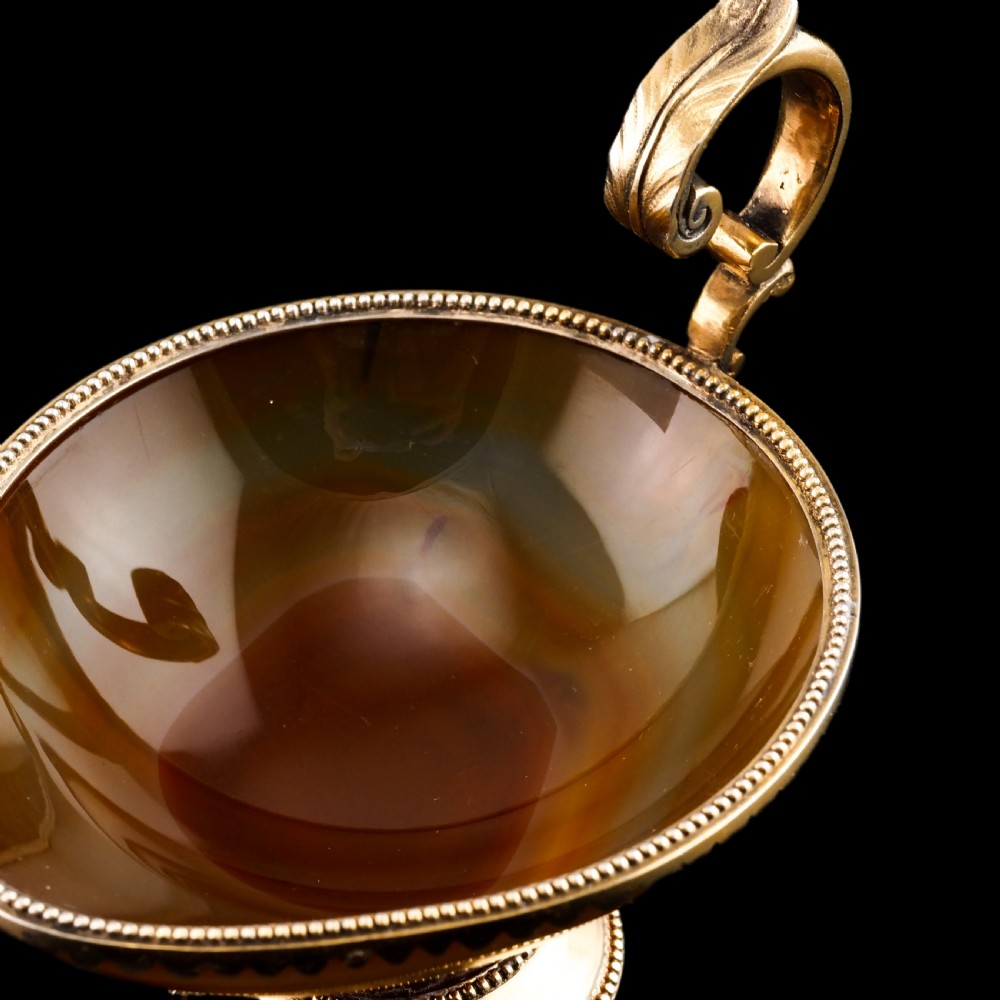 A 19th-century carved brown banded agate cup featuring a silver-gilt rim, pedestal base, and an ornate scroll handle. View XVI