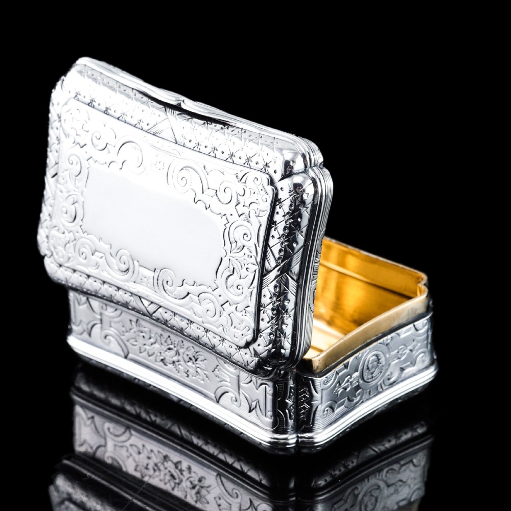 A Victorian sterling silver table snuff box with shaped cartouche outlines, foliage engraving, and an architectural scene. View XV