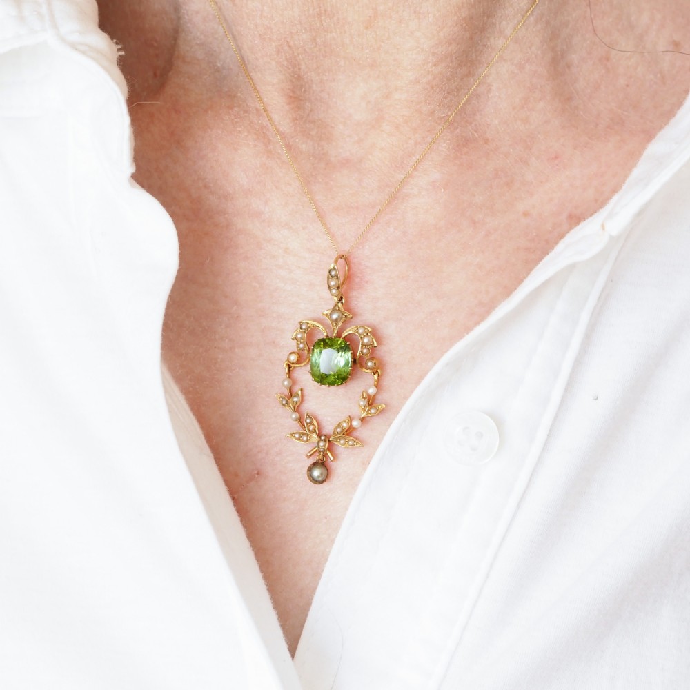 A late Victorian 14-carat yellow gold openwork pendant featuring a cushion-cut green peridot encircled by seed pearl foliate scrolls and a dangling pearl drop. View X