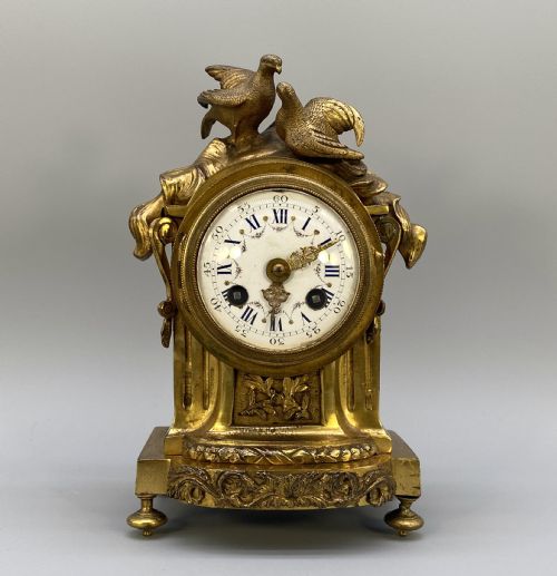 fig 1. An antique French gilt bronze mantel clock surmounted by two sculpted doves, featuring an ornate pedestal base and a white enamel dial with Roman numerals.