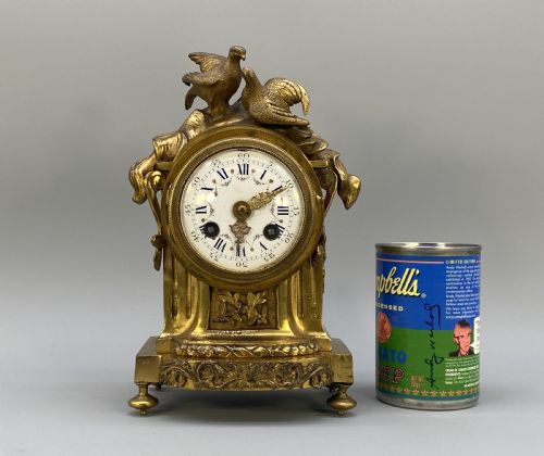 fig 2. An antique French gilt bronze mantel clock surmounted by two sculpted doves, featuring an ornate pedestal base and a white enamel dial with Roman numerals.