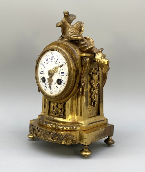 fig 3. An antique French gilt bronze mantel clock surmounted by two sculpted doves, featuring an ornate pedestal base and a white enamel dial with Roman numerals.