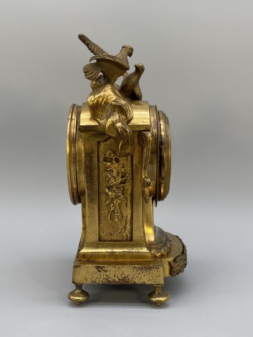fig 6. An antique French gilt bronze mantel clock surmounted by two sculpted doves, featuring an ornate pedestal base and a white enamel dial with Roman numerals.