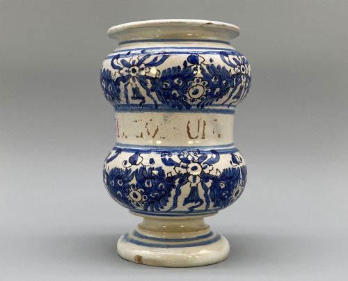 fig 2. An 18th-century Italian majolica albarello pharmacy jar featuring a waisted silhouette with hand-painted blue floral patterns and central apothecary inscription.