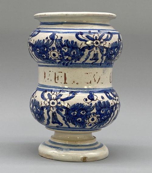 fig 3. An 18th-century Italian majolica albarello pharmacy jar featuring a waisted silhouette with hand-painted blue floral patterns and central apothecary inscription.