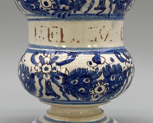 fig 5. An 18th-century Italian majolica albarello pharmacy jar featuring a waisted silhouette with hand-painted blue floral patterns and central apothecary inscription.