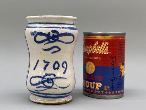 fig 2. A cylindrical tin-glazed earthenware albarello jar with a waisted body featuring hand-painted blue scrollwork and the date 1709 on a cream-white background.