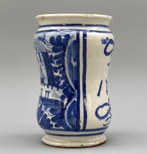 fig 3. A cylindrical tin-glazed earthenware albarello jar with a waisted body featuring hand-painted blue scrollwork and the date 1709 on a cream-white background.