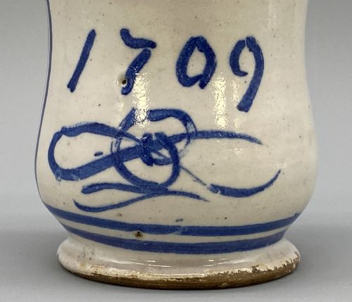 fig 4. A cylindrical tin-glazed earthenware albarello jar with a waisted body featuring hand-painted blue scrollwork and the date 1709 on a cream-white background.