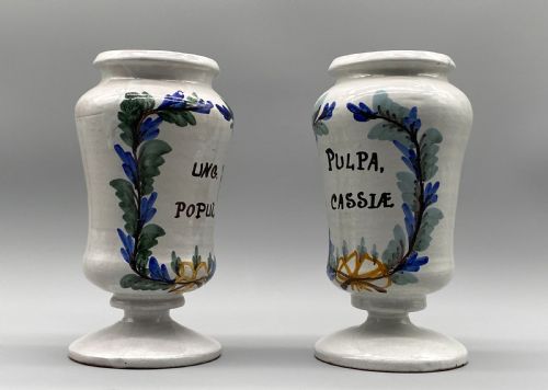 fig 3. A pair of white tin-glazed ceramic albarello apothecary jars featuring hand-painted green foliage wreaths and Latin script on pedestal bases.