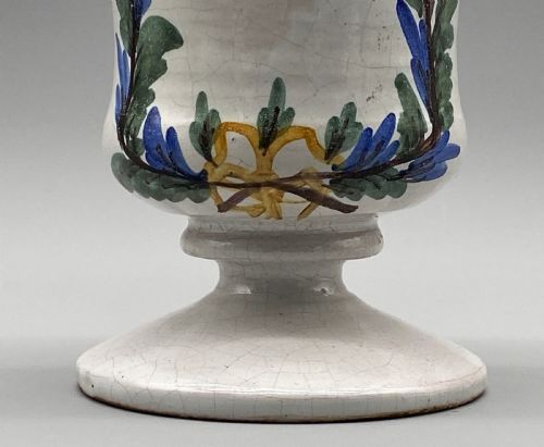 fig 7. A pair of white tin-glazed ceramic albarello apothecary jars featuring hand-painted green foliage wreaths and Latin script on pedestal bases.