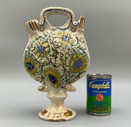 fig 1. An 18th-century Italian polychrome majolica pilgrim flask formed as a scallop shell with an overhead handle, side loops, and hand-painted floral motifs in blue, yellow, and green over a cream glazed surface.