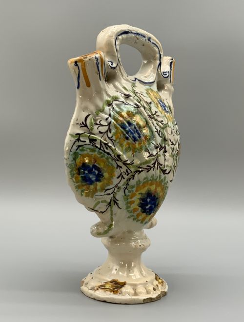 fig 2. An 18th-century Italian polychrome majolica pilgrim flask formed as a scallop shell with an overhead handle, side loops, and hand-painted floral motifs in blue, yellow, and green over a cream glazed surface.