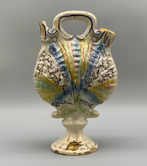 fig 5. An 18th-century Italian polychrome majolica pilgrim flask formed as a scallop shell with an overhead handle, side loops, and hand-painted floral motifs in blue, yellow, and green over a cream glazed surface.