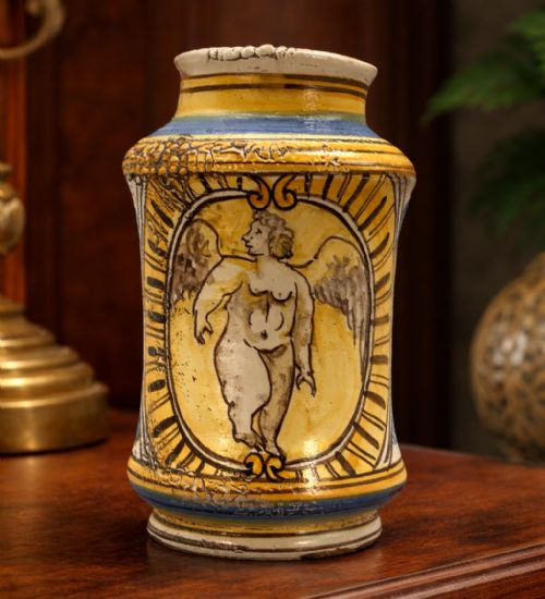 fig 1. A waisted cylindrical tin-glazed majolica albarello jar decorated in yellow, blue, and dark brown hand-painted pigments featuring a central winged figure motif.