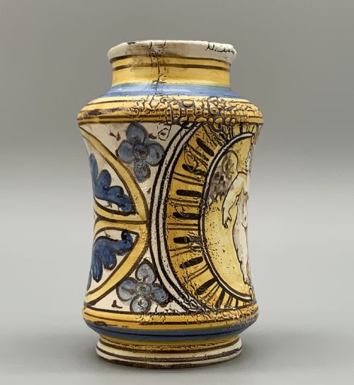 fig 3. A waisted cylindrical tin-glazed majolica albarello jar decorated in yellow, blue, and dark brown hand-painted pigments featuring a central winged figure motif.