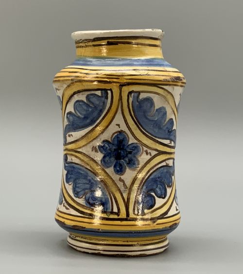 fig 4. A waisted cylindrical tin-glazed majolica albarello jar decorated in yellow, blue, and dark brown hand-painted pigments featuring a central winged figure motif.