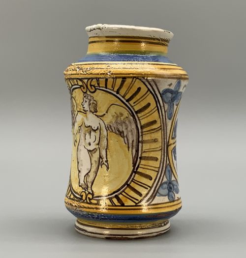 fig 5. A waisted cylindrical tin-glazed majolica albarello jar decorated in yellow, blue, and dark brown hand-painted pigments featuring a central winged figure motif.
