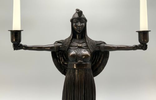 fig 2. A patinated bronze Egyptian Revival candelabra featuring a female figure with outstretched arms holding two candle sockets, standing on a decorated triangular base.