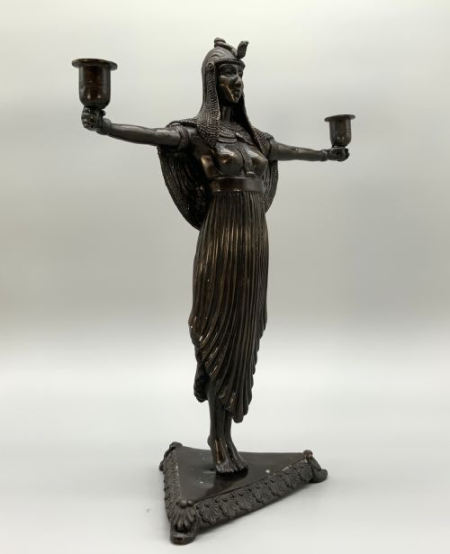 fig 3. A patinated bronze Egyptian Revival candelabra featuring a female figure with outstretched arms holding two candle sockets, standing on a decorated triangular base.