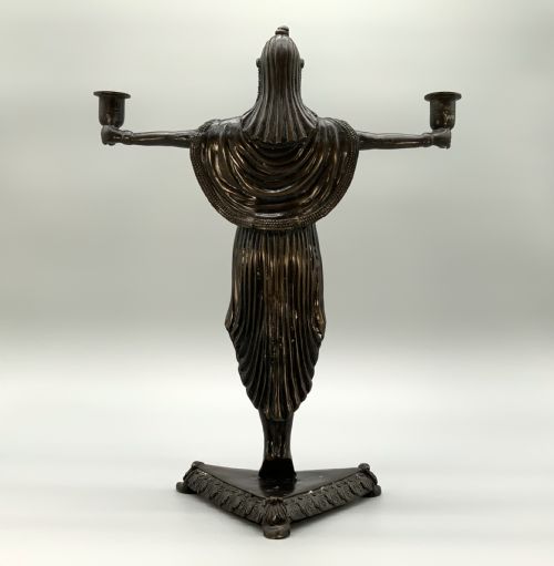 fig 5. A patinated bronze Egyptian Revival candelabra featuring a female figure with outstretched arms holding two candle sockets, standing on a decorated triangular base.