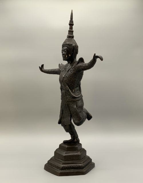 fig 3. A dark bronze sculpture depicting a classical court dancer in ceremonial attire with a tall spired headdress, balanced on one foot atop an octagonal stepped base.
